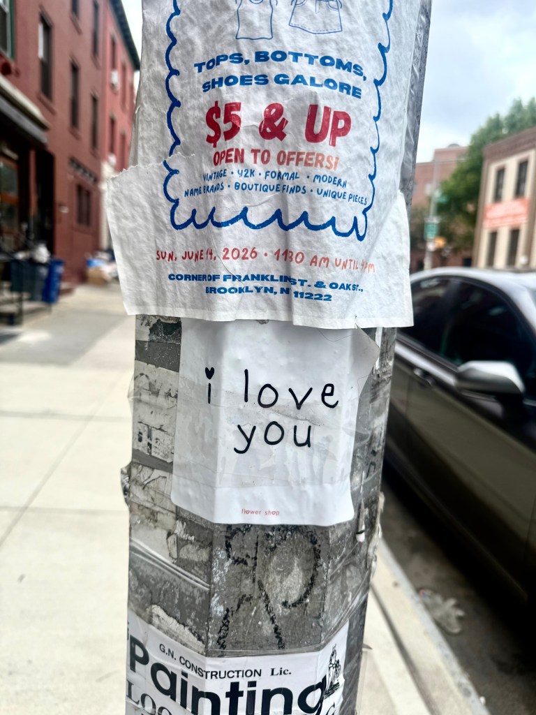 A street pole covered with various flyers, including one advertising a vintage clothing sale and a handwritten note that says 'i love you'.