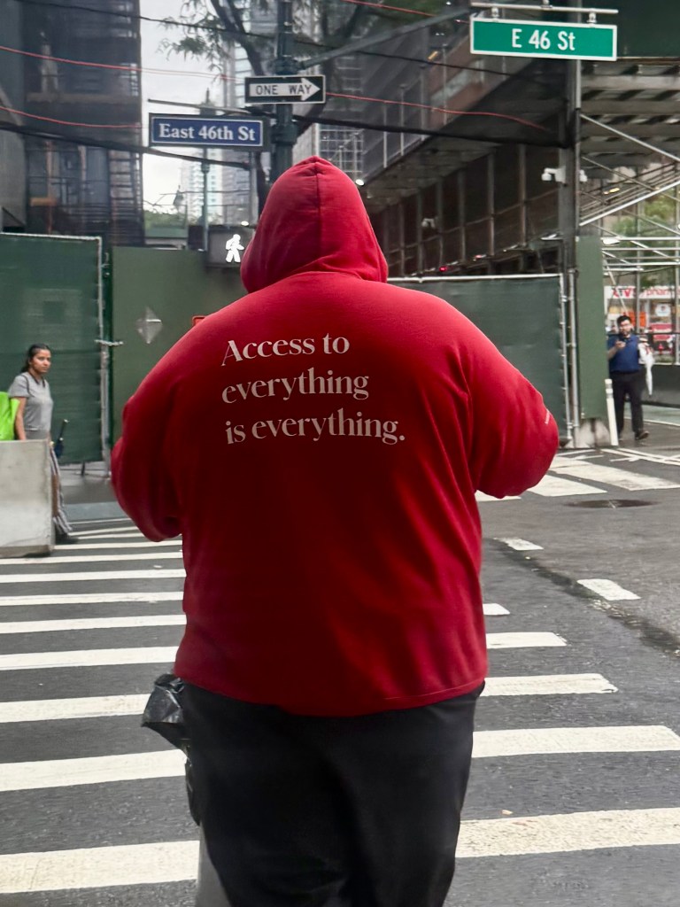 A person wearing a red hoodie with the text 'Access to everything is everything' printed on the back, standing on a crosswalk with street signs in the background.