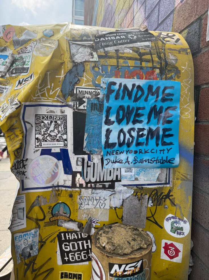 A streetpost covered in various stickers and graffiti, featuring a prominent blue sign that reads 'FIND ME LOVE ME LOSE ME' along with 'NEWYORKCITY' and 'DUKE A BARNSTABLE.'