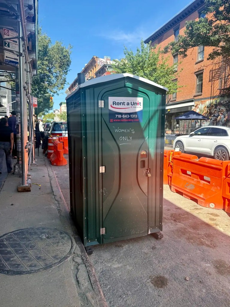 A green portable restroom labeled 'Women's Only' with a sign for 'Rent a Unit' in a urban street setting with construction barriers in the background.