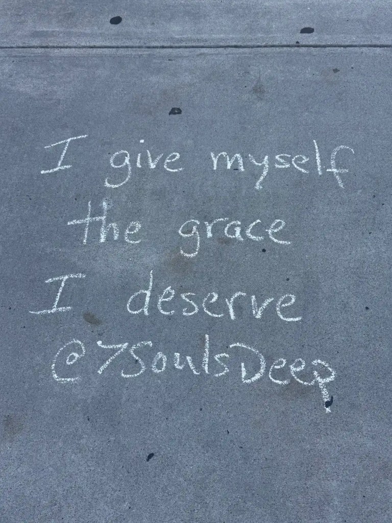 Chalk writing on pavement that reads 'I give myself the grace I deserve @7SoulsDeep'.