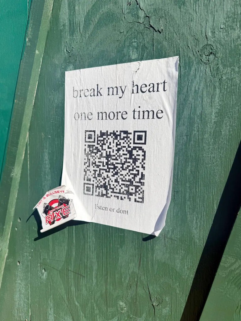 A poster on a green wooden surface with the text 'break my heart one more time' and a QR code beneath it, accompanied by an AXE label.