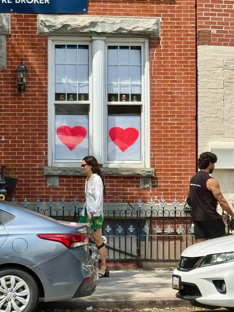 A woman in a white top and green shorts walks past a building with two windows displaying red heart decorations. A man in a black tank top is seen walking nearby, while parked cars are visible in the foreground.