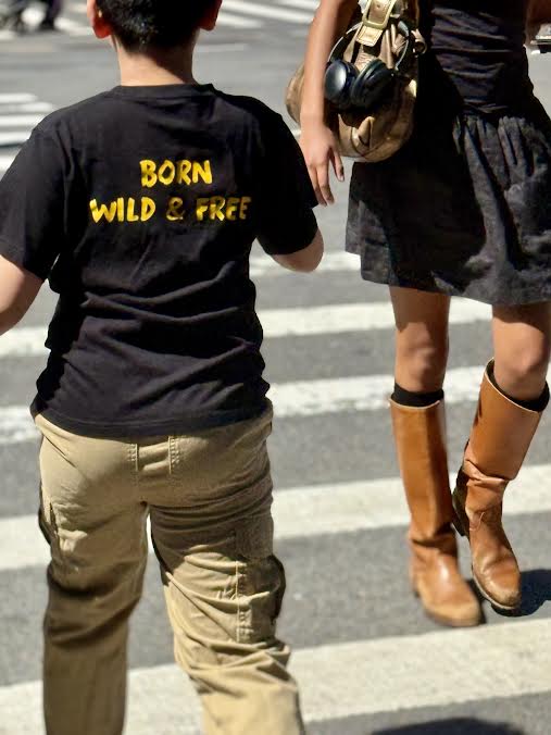 A child wearing a black t-shirt that says 'BORN WILD & FREE' walks in a crosswalk alongside an adult woman wearing a dark dress and brown knee-high boots.