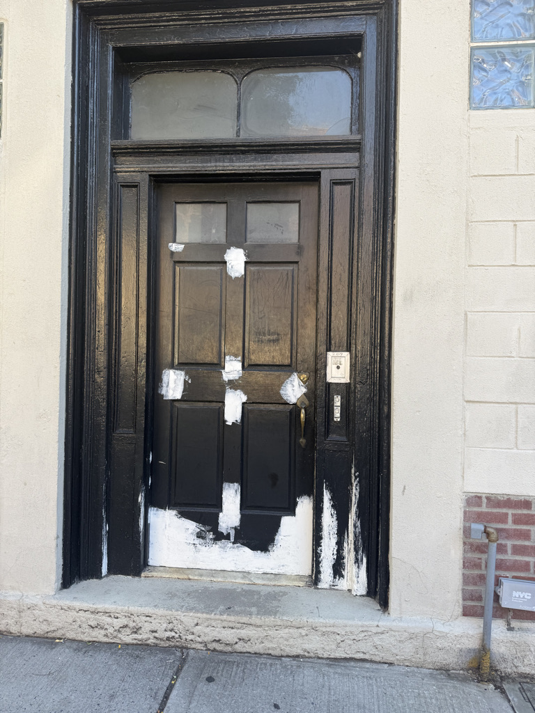 A weathered black door with visible paint damage and semi-peeled paint, situated on a sidewalk next to a brick wall.