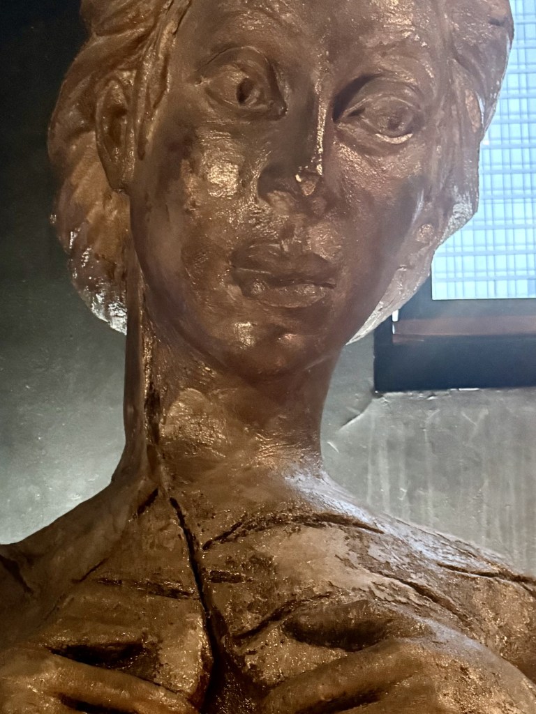 A close-up of a large, dark sculpture depicting a woman's head and upper torso, featuring detailed facial expressions and textured hair.