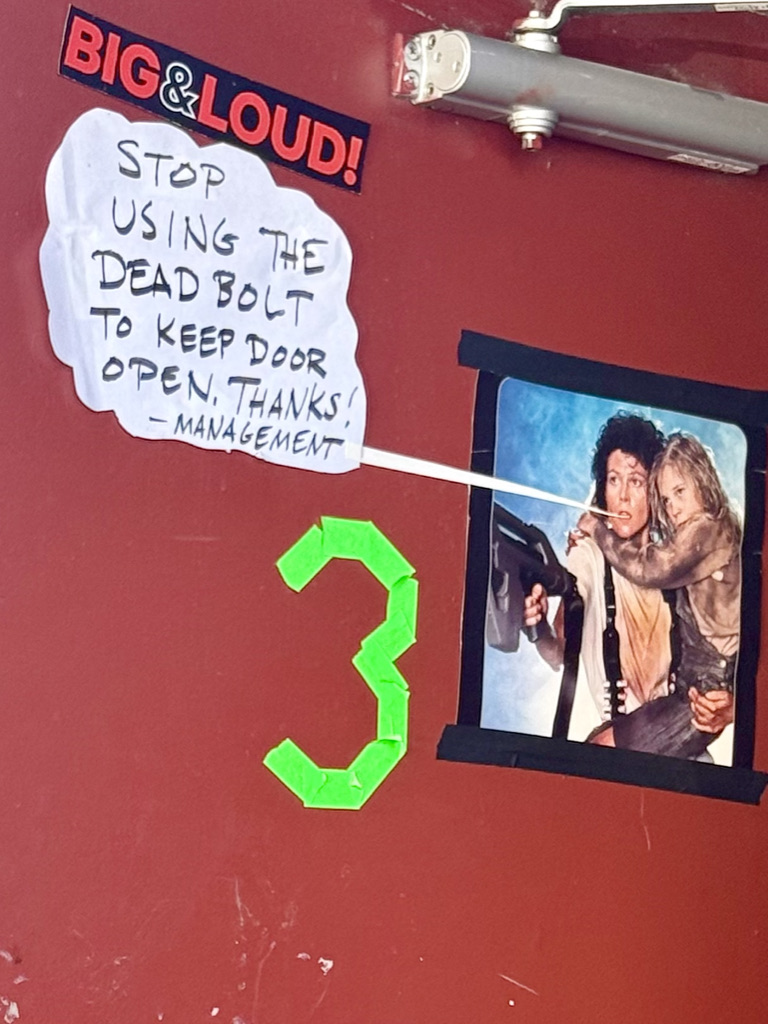 A red door displaying a sign instructing not to use the deadbolt to keep it open, with a note from management. A large green number '3' is taped to the door, and a photo of two individuals, one holding a gun, is also attached.