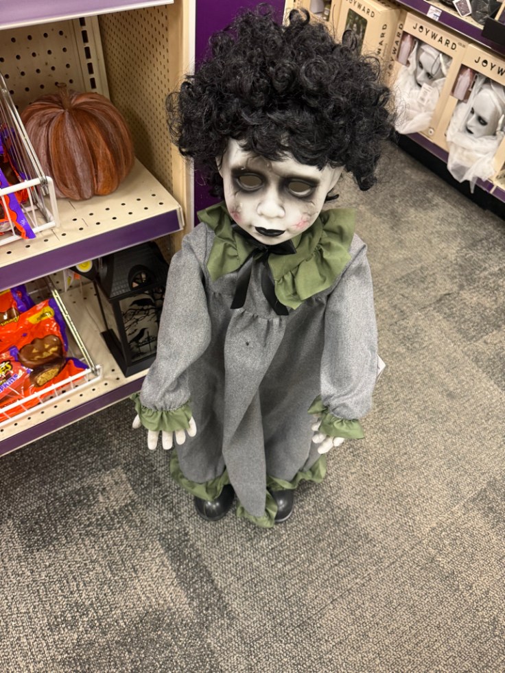 A spooky clown doll with curly black hair, pale skin, and dark eyes, wearing a gray outfit with a green collar and cuffs, standing on a store shelf among Halloween decorations.