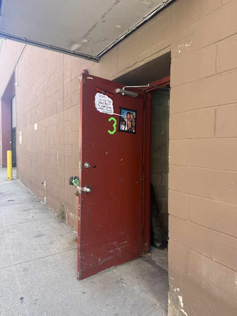 An open red door with a sign that reads 'BIG & LOUD!' and a photo attached, leading into a dimly lit area beside a brick wall.