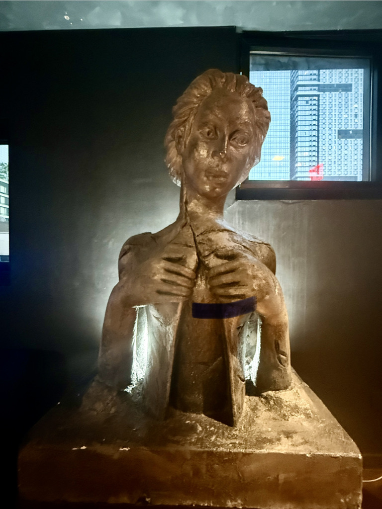 A bronze sculpture of a woman with an expressive face, holding her hands to her chest, set against a dark background with a window showcasing a cityscape.