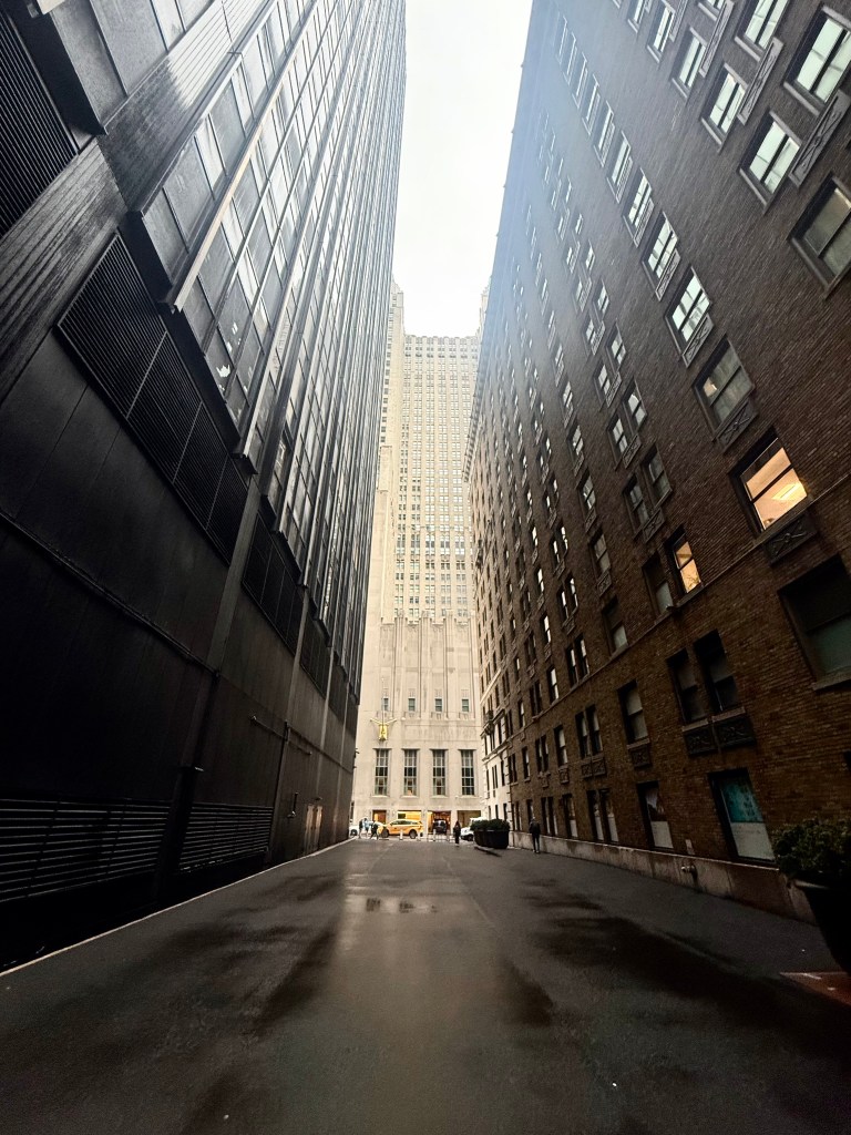 Narrow urban alleyway flanked by tall buildings, with a clear sky above and a street visible in the background.