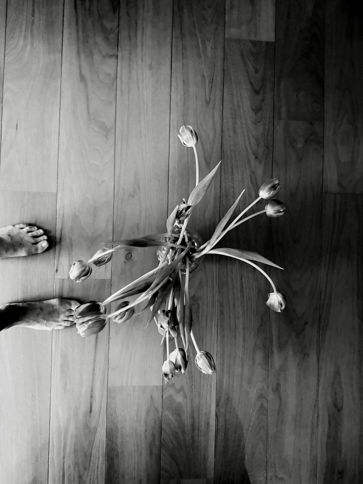 A black and white photo of a bouquet of tulips on a wooden floor, with a person's bare feet visible at the bottom of the image.