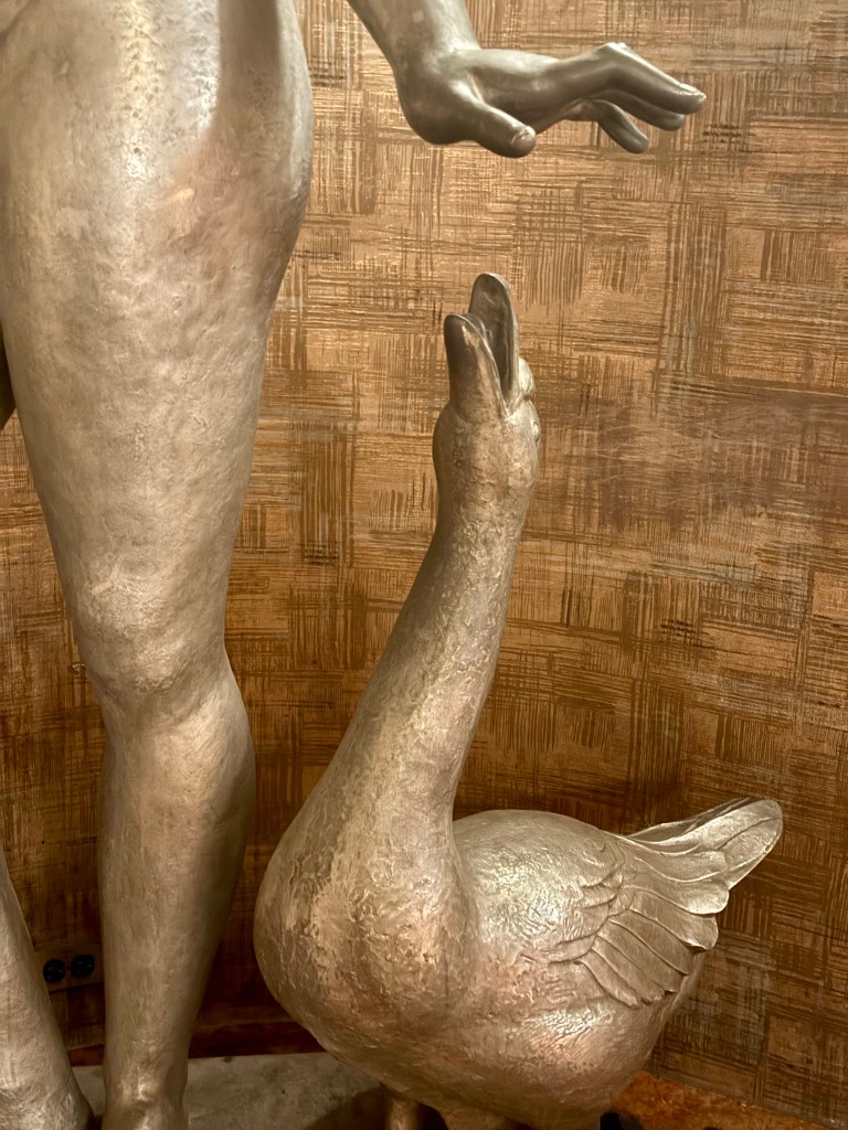 A close-up of a sculpture featuring a stylized figure of a person and a goose, highlighting the detailed textures of both figures.
