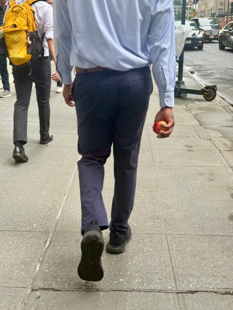 A man wearing a blue checkered shirt walks down a city sidewalk holding a red apple in his right hand.
