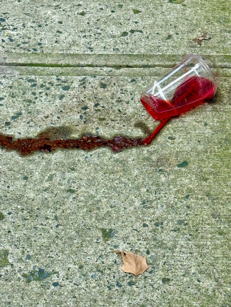 A spilled plastic cup with red liquid on a textured concrete surface, with a dry leaf nearby.