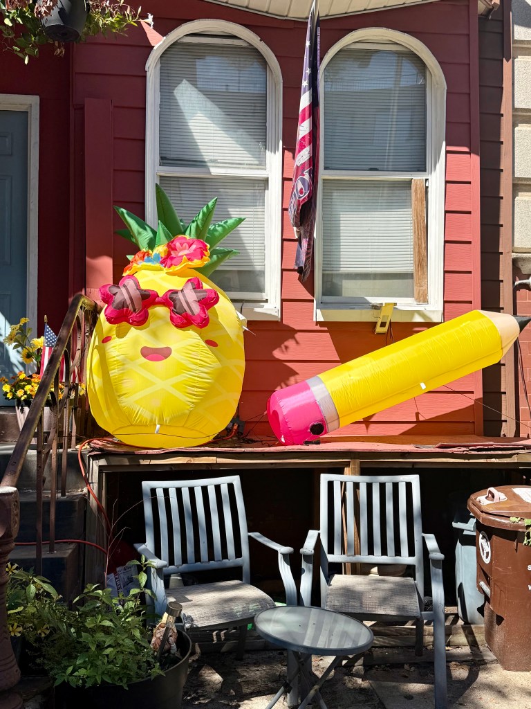 A colorful inflatable pineapple with sunglasses and a floral crown, alongside a large inflatable pencil, positioned in front of a red house with two windows and an American flag.