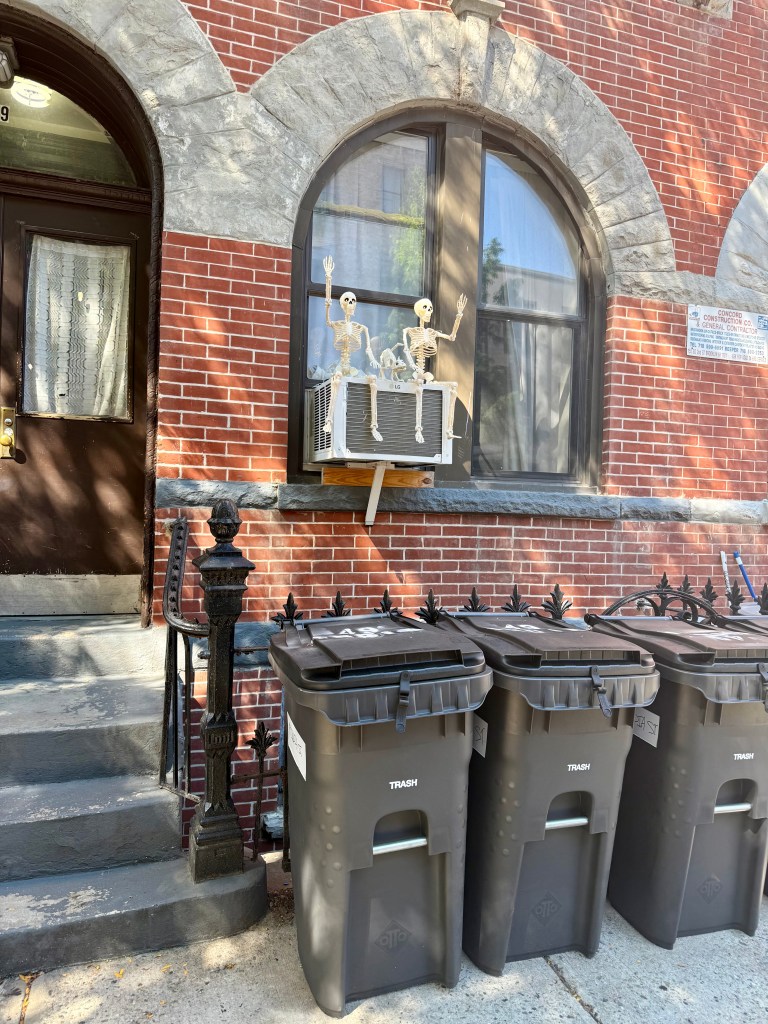 Two skeleton decorations sitting on an air conditioning unit in a window, with a brick building entrance and dark trash bins lined up below.