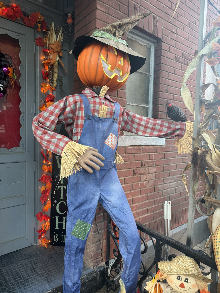 A whimsical scarecrow with a pumpkin head and a smiling face, wearing a plaid shirt and denim overalls, stands outside a decorated house. The scarecrow holds a small black bird and is surrounded by autumn decorations.