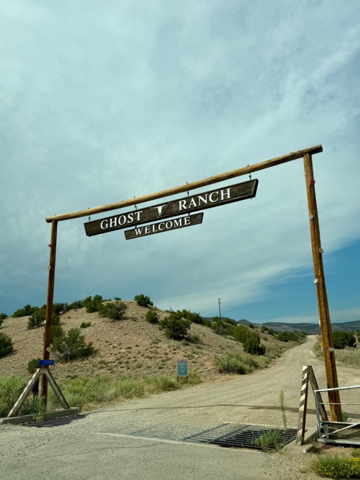 Welcome sign for Ghost Ranch, situated on a dirt road surrounded by hills and greenery under a cloudy sky.