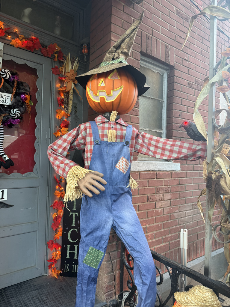 A scarecrow decoration with a pumpkin head and a straw hat, wearing a checkered shirt and denim overalls, standing outside a house adorned with autumn decorations.