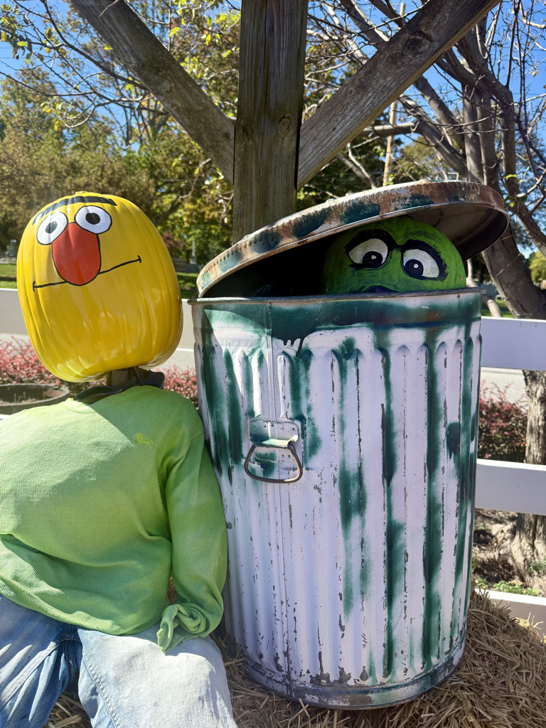 A decorative scene featuring a scarecrow on the left with a yellow painted pumpkin head and green shirt, sitting next to a colorful trash can with a green character peeking out.