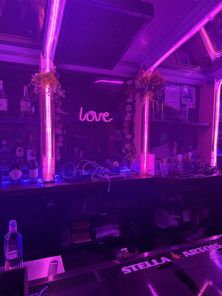 A brightly lit bar with purple neon lights, featuring a sign that says 'love' and decorated with flowers and bottles on the shelf.