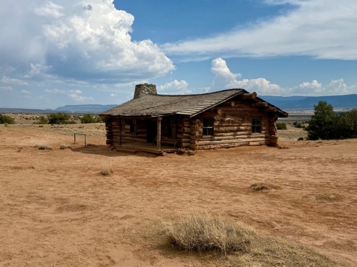 A rustic log cabin surrounded by a sandy landscape under a partly cloudy sky.