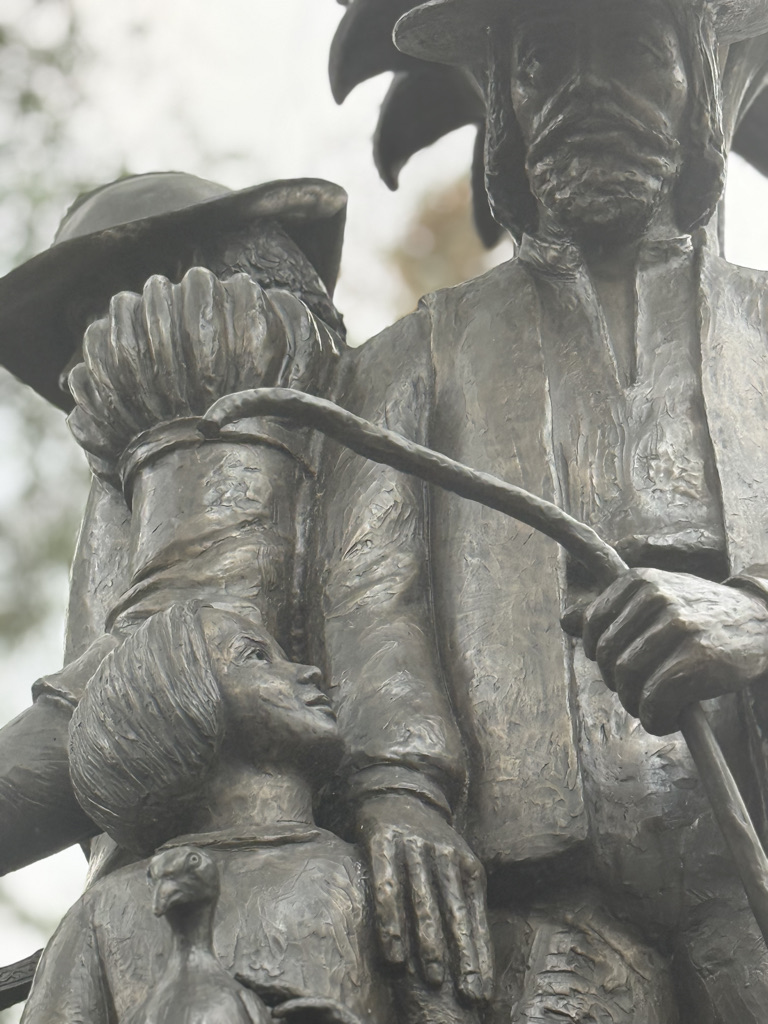 Close-up of a bronze statue depicting a farmer holding a stick, standing alongside a child looking up, with another figure partially visible in the background.