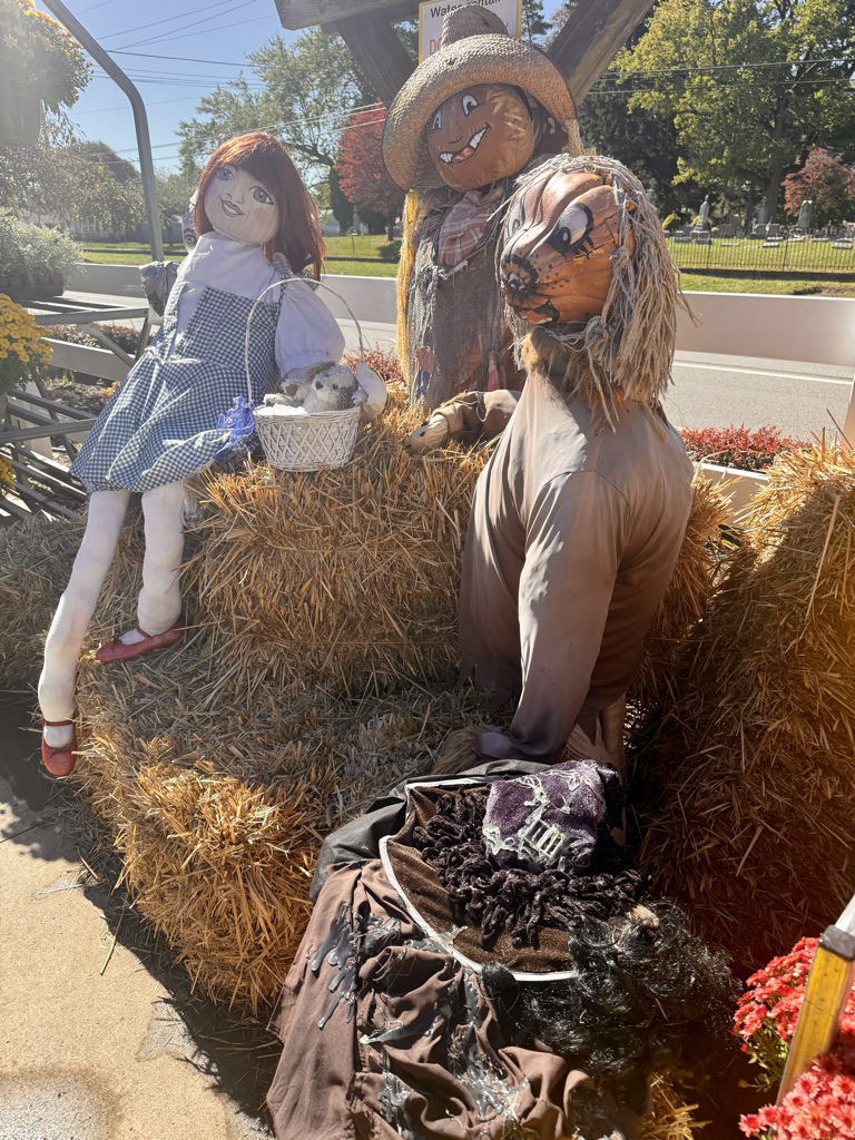 A whimsical autumn display featuring a doll in a blue dress and red shoes sitting on hay bales, alongside a scarecrow with a smiling pumpkin head and a lion figure dressed in ragged clothing.