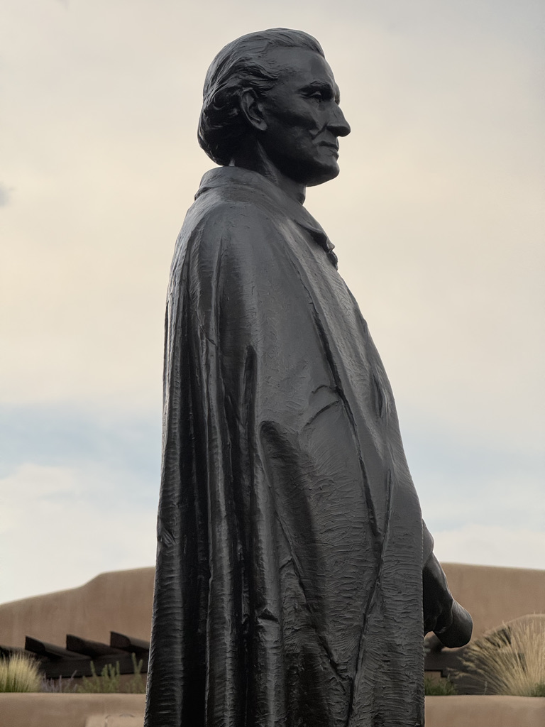 A profile view of a bronze statue of a historical figure wearing a cape, set against a cloudy sky.