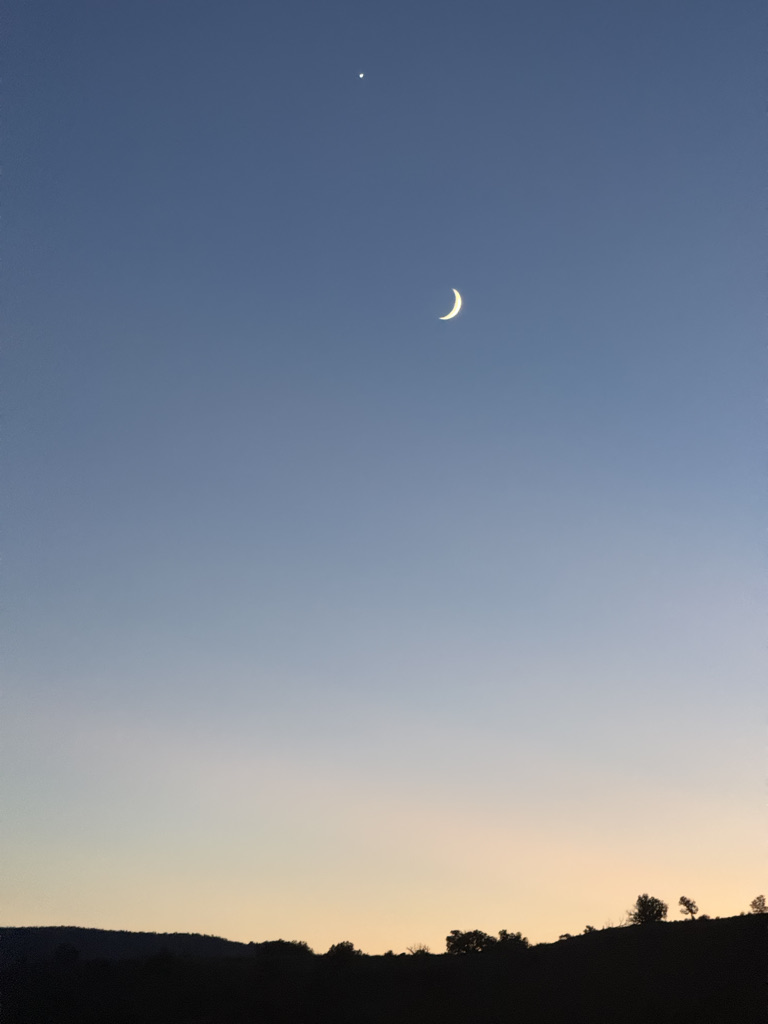 A crescent moon and a bright star visible in a clear evening sky with soft gradients of blue and orange at the horizon.