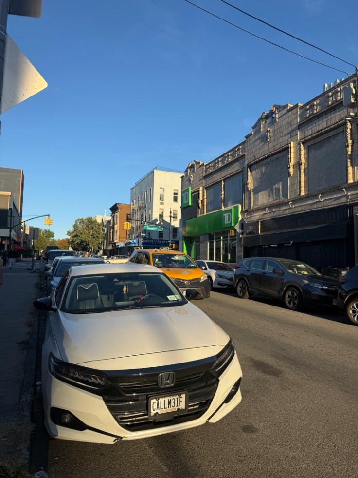 Busy street scene featuring a white Honda parked on the left, with various vehicles, shops, and a clear blue sky in the background.