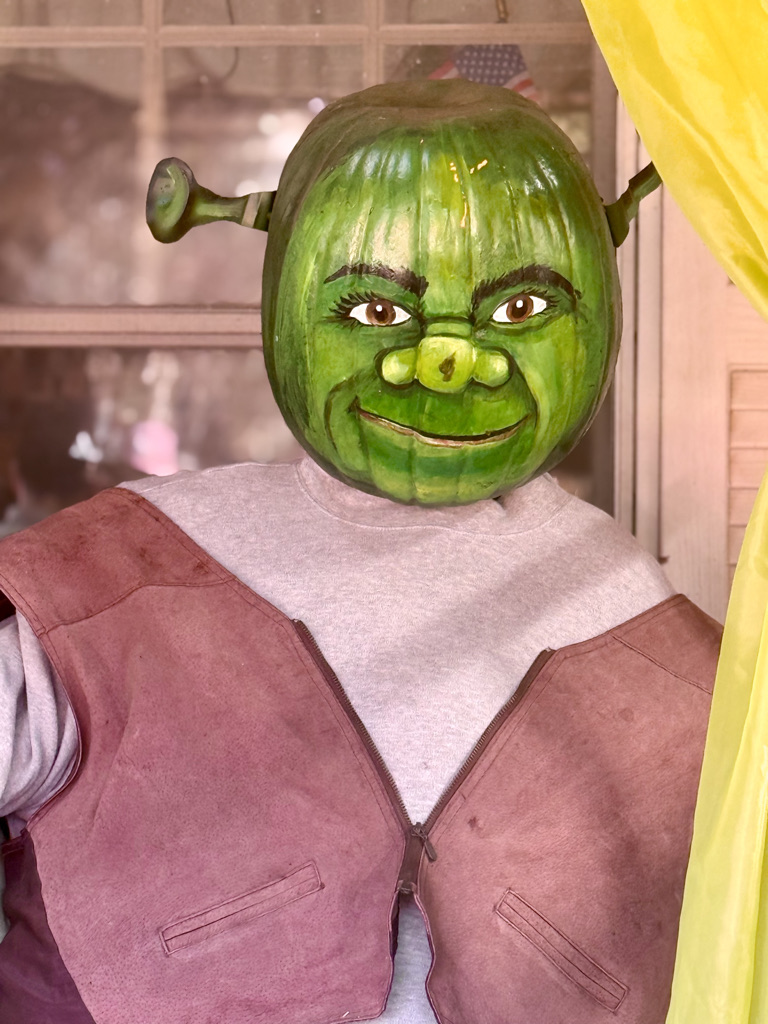 A person dressed as a character resembling Shrek, featuring a green pumpkin head painted with facial details, wearing a gray shirt and a brown vest, standing in front of a window.