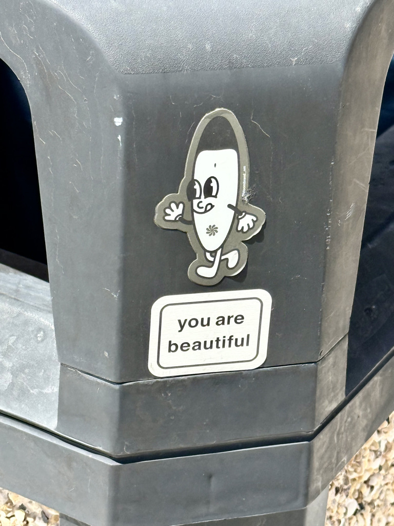 A black waste bin featuring a cartoon character sticker with a smiling face and arms, alongside a text sticker that reads 'you are beautiful'.