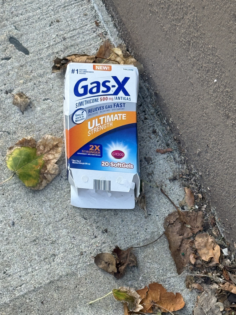 An empty package of Gas-X Ultimate Strength, a medication for gas relief, lying on a sidewalk surrounded by fallen leaves.