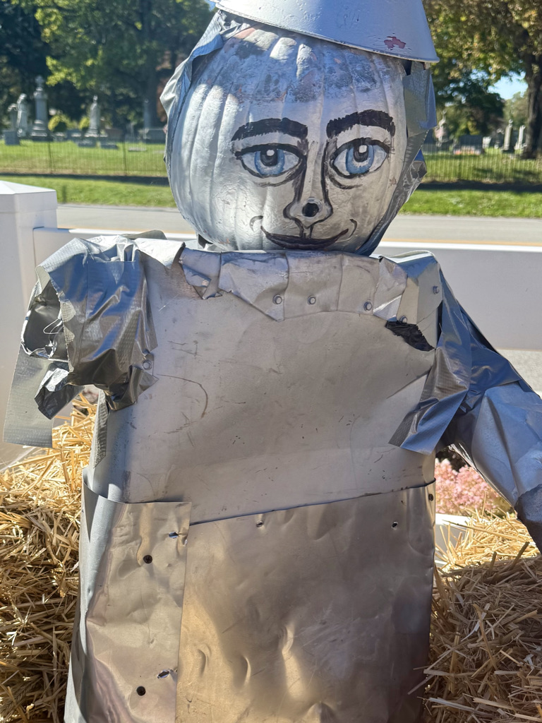 A scarecrow made from a pumpkin head covered in aluminum foil and a silver outfit, set against a backdrop of trees and a cemetery.