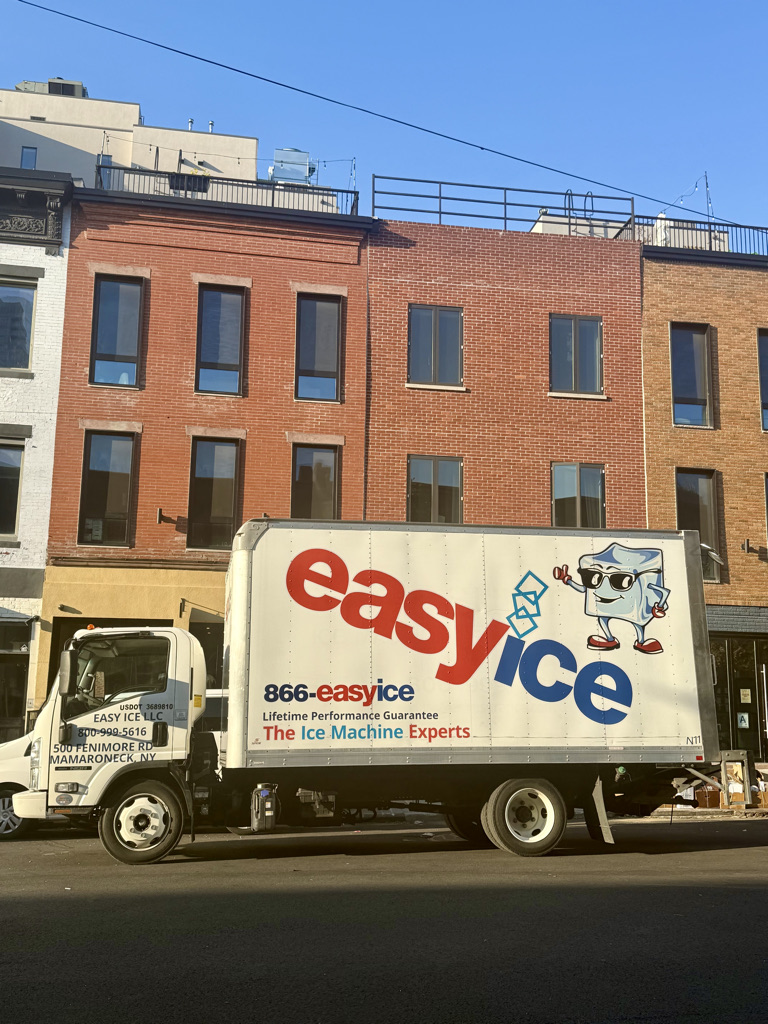 A delivery truck for Easy Ice parked on a city street, featuring a large logo with a cartoon ice cube mascot and the text '866-easyice' and 'Lifetime Performance Guarantee'. The background includes brick buildings and a clear blue sky.