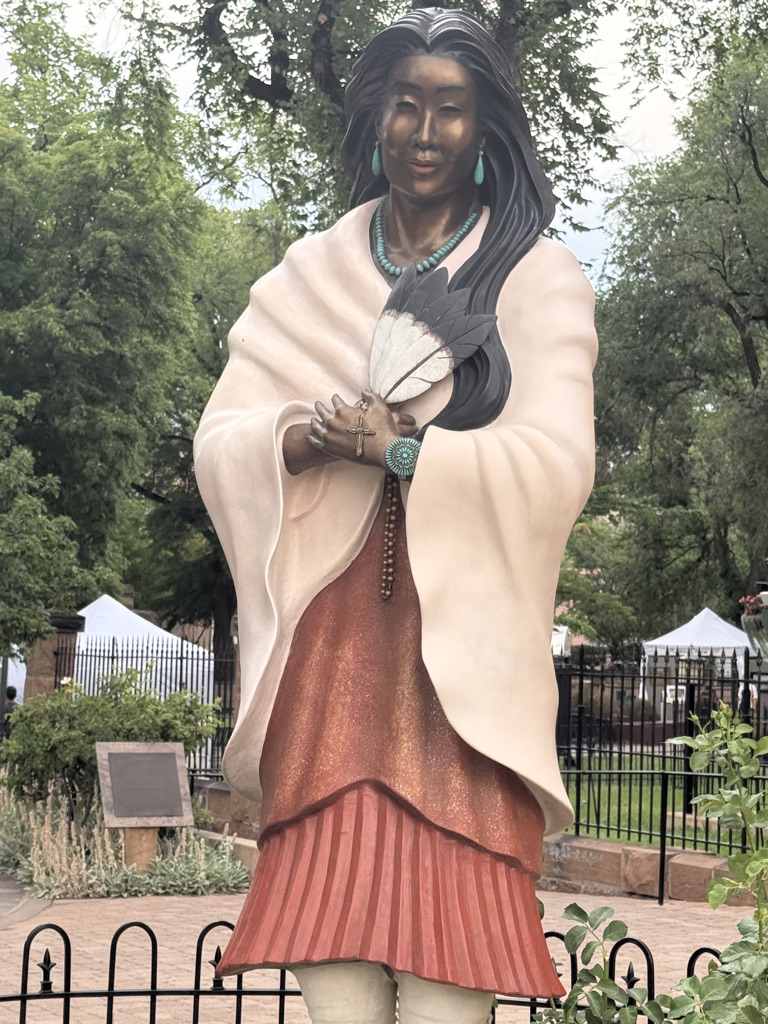 A statue of a woman with long dark hair, wearing a traditional outfit adorned with turquoise jewelry and holding a feather fan, positioned in a green outdoor setting.