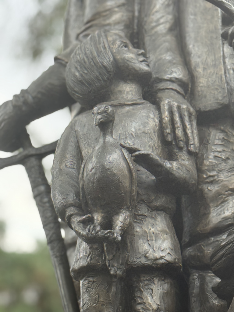Close-up of a bronze sculpture depicting a child holding a small bird, with an adult figure beside them.