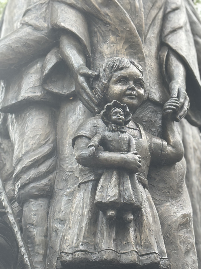 Close-up of a bronze sculpture depicting a child holding a doll, alongside an adult figure.