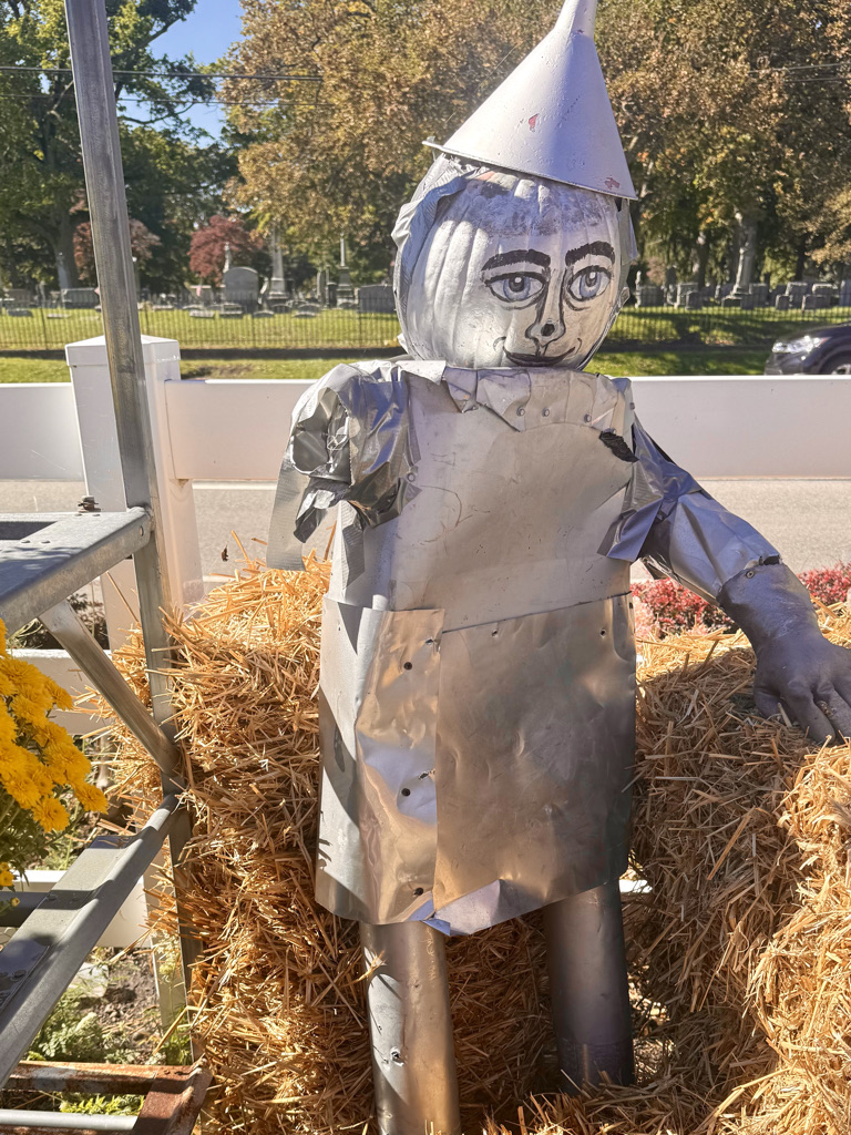A whimsical scarecrow figure made of metal and painted to resemble a character, standing on a bed of straw with colorful flowers nearby.
