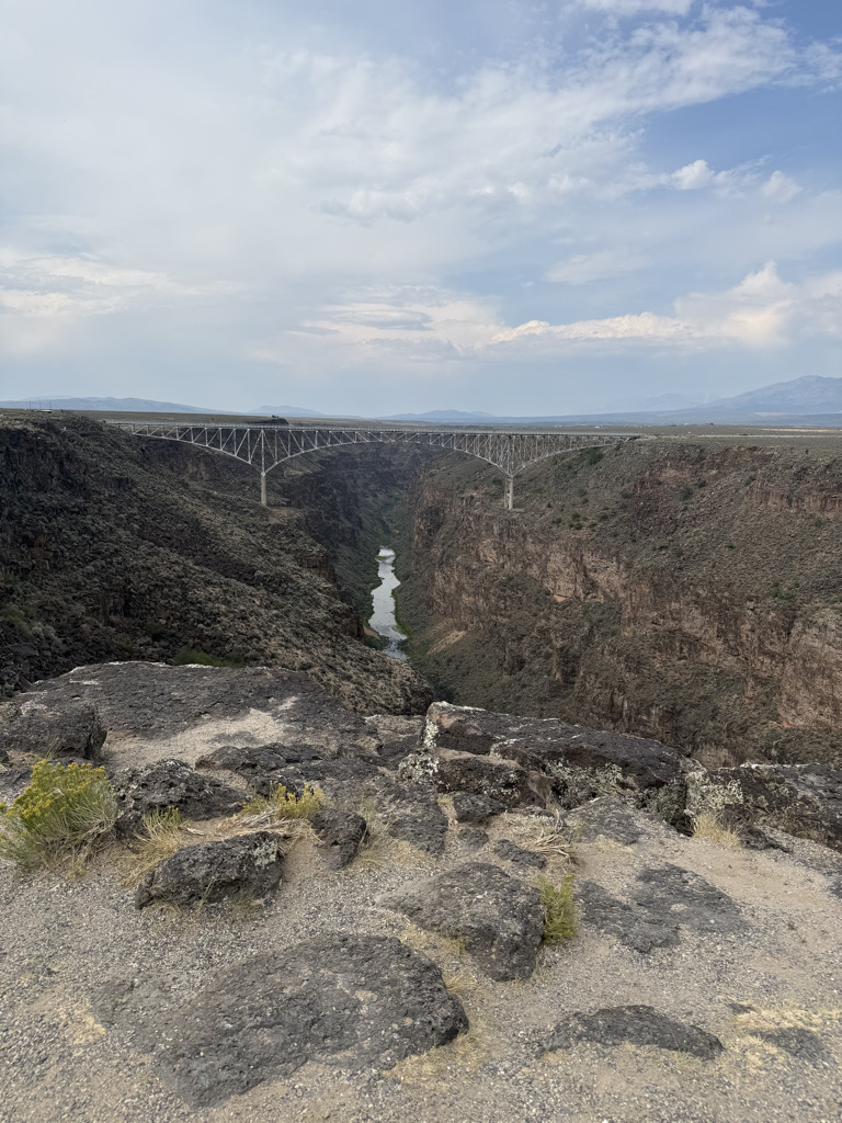 A view of a deep canyon with a river flowing through it, featuring a large steel bridge spanning across the canyon. The landscape is rocky with sparse vegetation under a cloudy sky.
