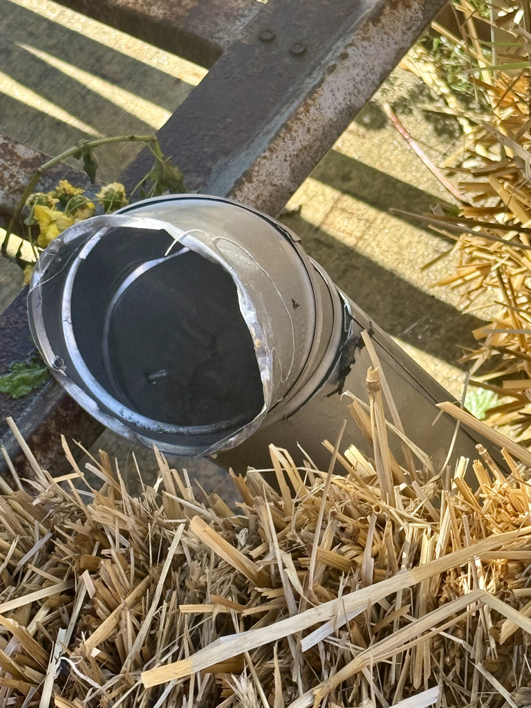 A broken metal pipe resting on a bed of straw, with sunlight reflecting off its surface.