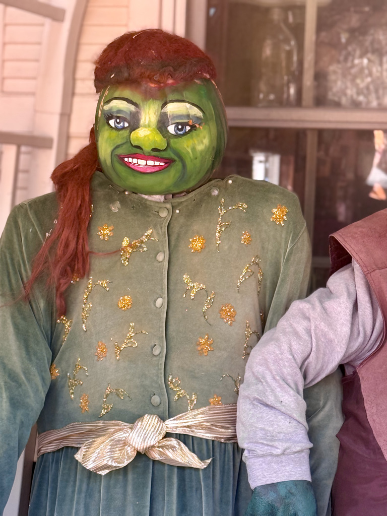 A whimsical character dressed in a velvet green outfit with floral embellishments, featuring a painted green face, bright red hair, and a cheerful smile.