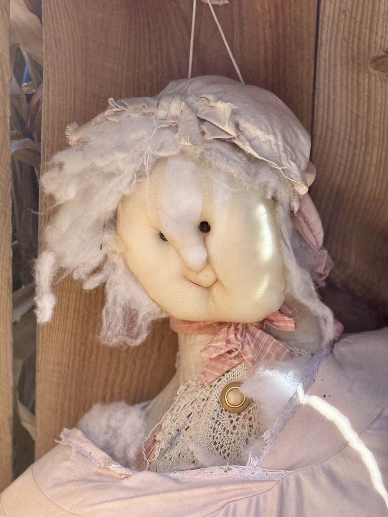 Close-up of a handmade doll with a soft, smiling face, wearing a pink dress and a matching cap, adorned with frayed fabric and cotton-like hair.