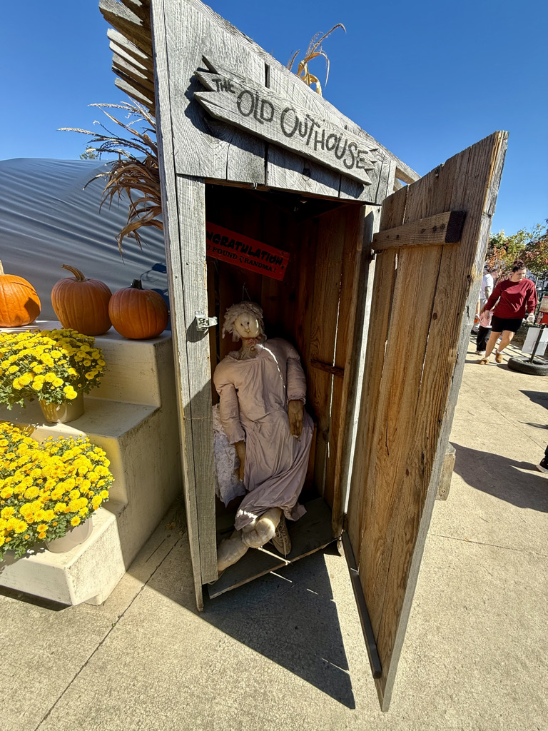 A wooden outhouse with the sign 'The Old Outhouse,' featuring a decorative, lifelike figure dressed in a long dress seated inside. Bright yellow flowers and pumpkins are in the foreground, with a clear blue sky overhead.