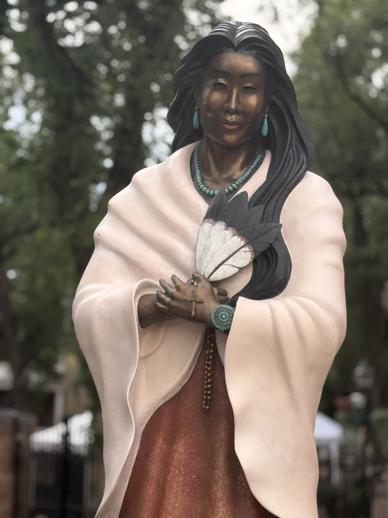 Statue of a woman wearing a white shawl, holding a fan, adorned with jewelry, with a serene expression and long hair, set in a natural outdoor environment.