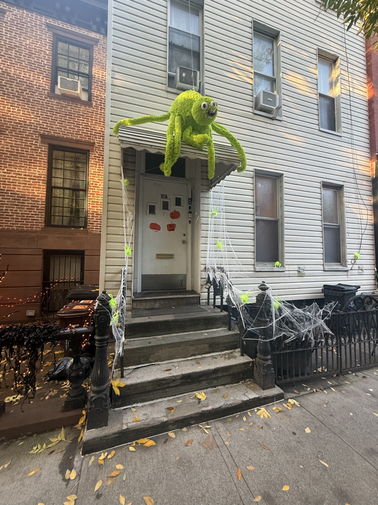 A green, plush spider decoration is perched on the entrance of a house, surrounded by Halloween decorations, including fake spider webs and leaves scattered on the ground.