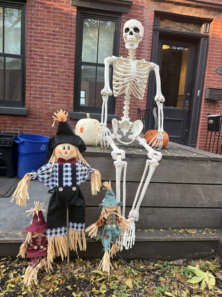 A Halloween decoration scene featuring a skeleton sitting beside a colorful scarecrow and two smaller scarecrow figures, with pumpkins and a brick wall in the background.
