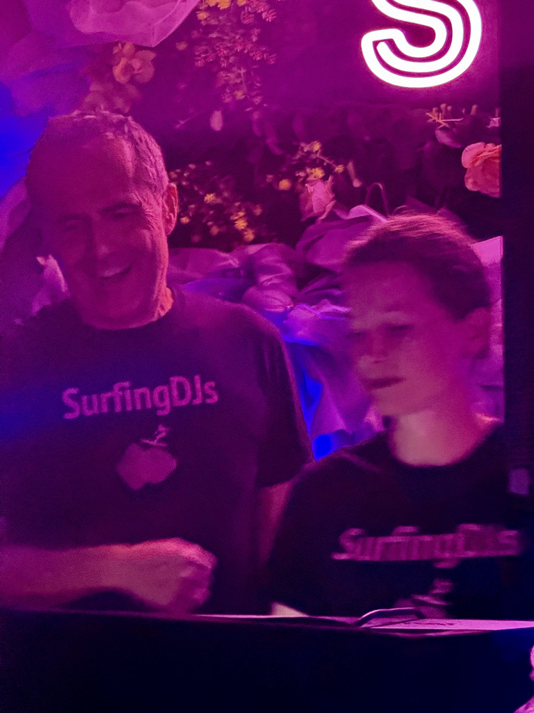 Two individuals smiling and interacting at a music event, both wearing black shirts with 'SurfingDJs' printed on them, under purple and pink lighting.